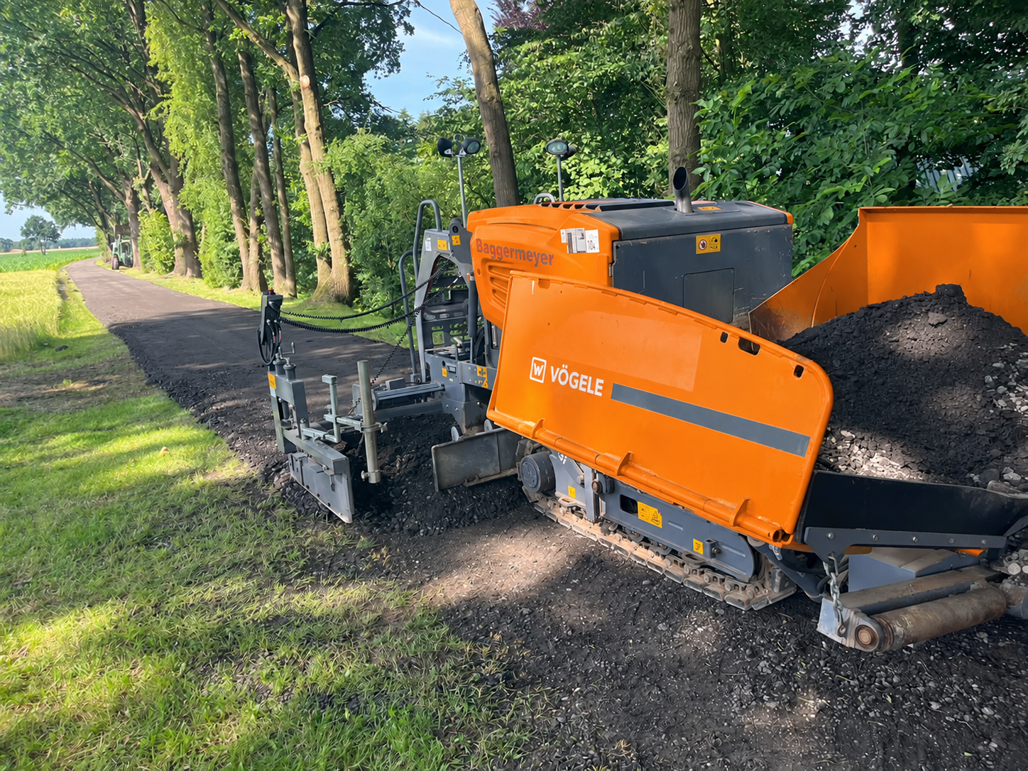 Orangefarbener Asphaltfertiger auf einem Weg zwischen Bäumen